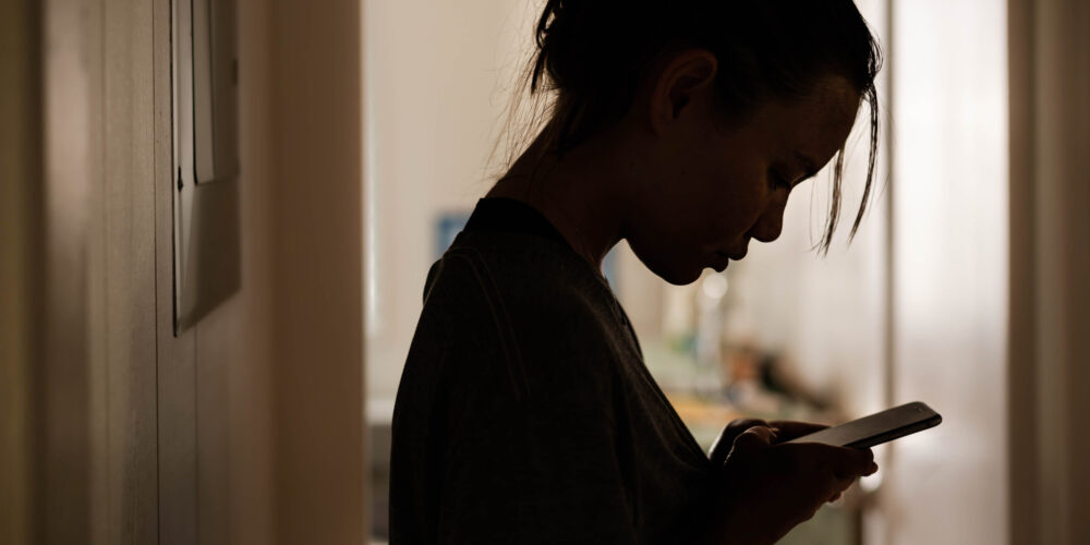 Silhouette of an upset-looking young woman viewing a message on her phone.