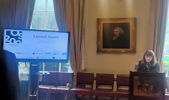 Dr Catherine Porter standing at a podium speaking into a microphone. A display PPT is on a monitor to her left